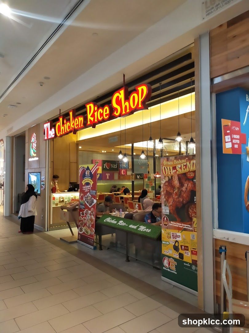 The exterior of a restaurant called "The Chicken Rice Shop" with a prominent red and yellow sign. Inside, customers are seated at tables, and staff can be seen behind the counter. Various promotional posters and banners are displayed outside the shop.
