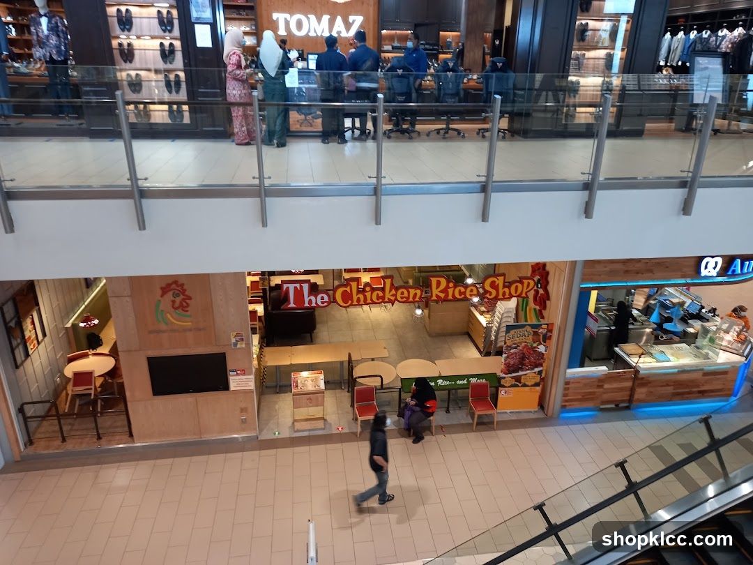 The exterior of a restaurant called "The Chicken Rice Shop" is visible. The shop has a large sign with the name and a chicken logo. Inside, tables and chairs are set up for dining. To the right of the chicken rice shop is another establishment with a blue and white sign that reads "Au".