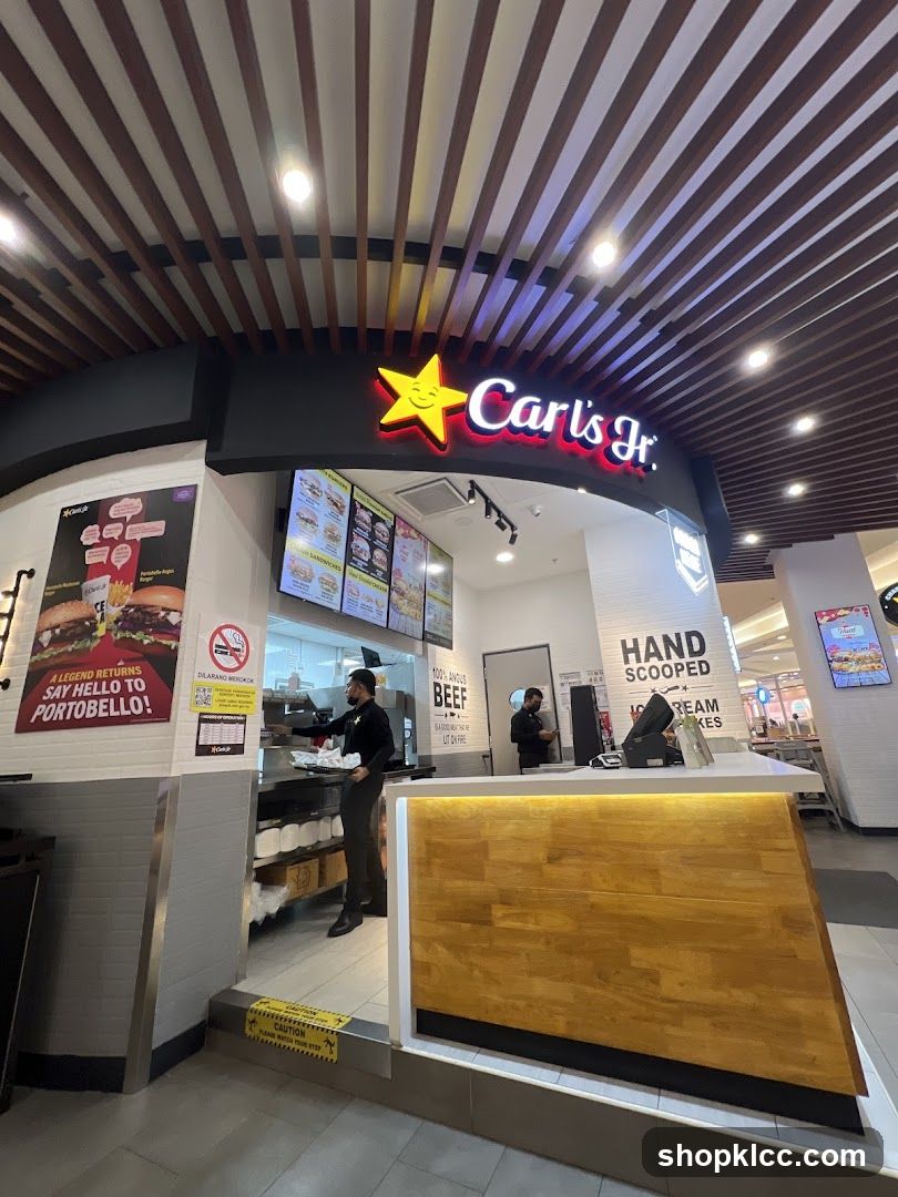 The exterior of a Carl's Jr. fast-food restaurant. The prominent Carl's Jr. logo with a star is visible above the entrance. Inside, employees are seen preparing food behind the counter. Menu boards displaying food items are mounted on the wall. A promotional poster for a Portobello burger is displayed to the left. The counter area features wooden paneling and a white countertop.