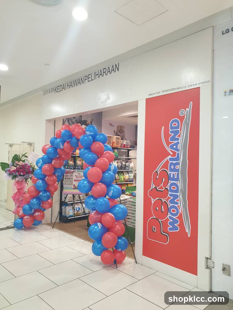 The exterior of a pet store with a red door featuring the "Pets Wonderland" logo. A balloon arch in red and blue decorates the entrance. The store name "KEDAI HAIWAN PELIHARAAN" is visible above the entrance. Shelves stocked with pet supplies are visible inside.