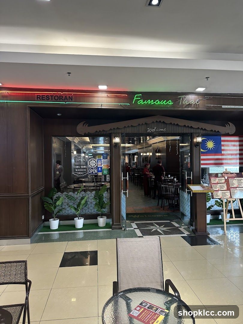The exterior of a Thai restaurant with neon signs reading "RESTORAN" and "Famous Thai". The entrance is framed by dark wood and features a "Welcome" mat. Inside, tables and chairs are visible, along with staff members. A Malaysian flag is displayed to the right of the entrance. A menu board is also visible outside the restaurant.