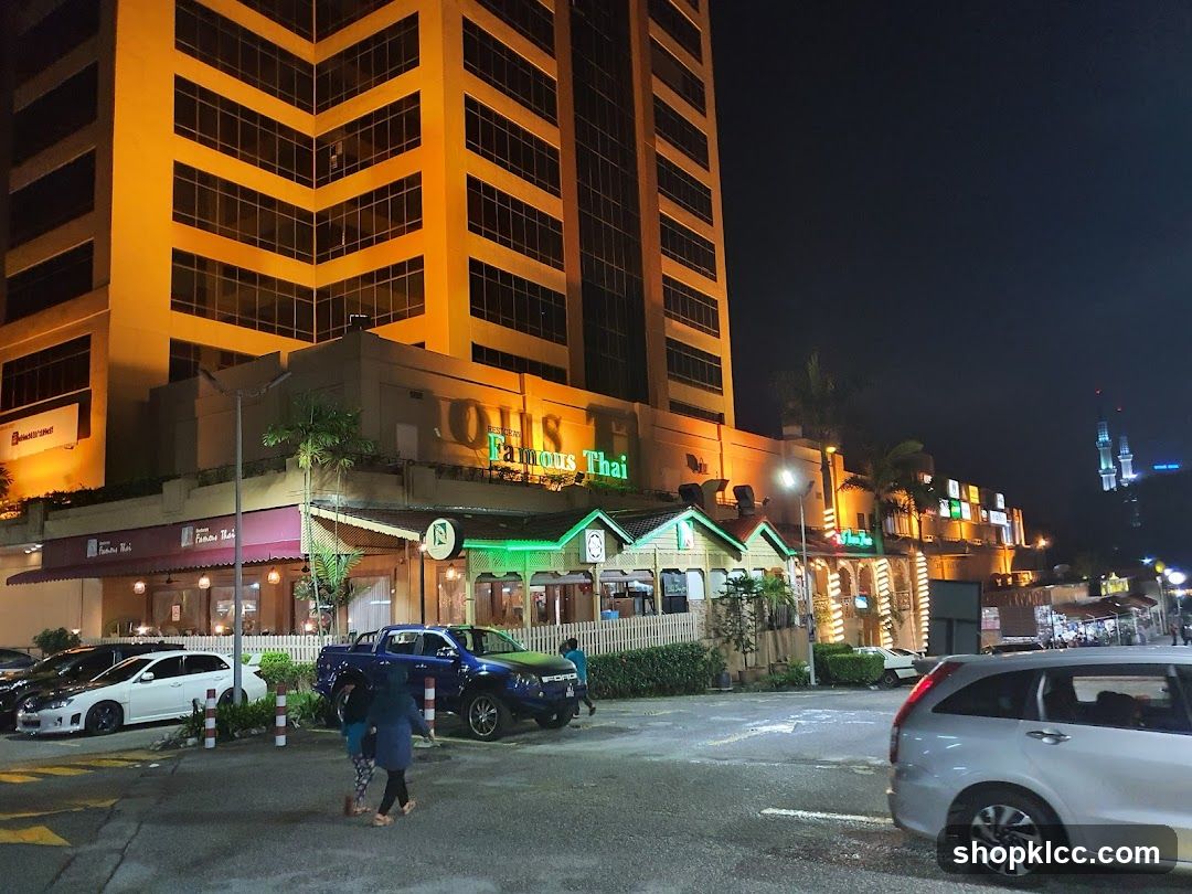 A night view of the exterior of a building with multiple businesses. The prominent business in the foreground is "Famous Thai" restaurant, with its name clearly visible on the facade. The restaurant has a traditional Thai architectural style with green accents and white fencing. Other businesses are visible further down the street, illuminated by various signs and lights. Cars are parked in front of the establishments, and a few people are walking on the sidewalk.