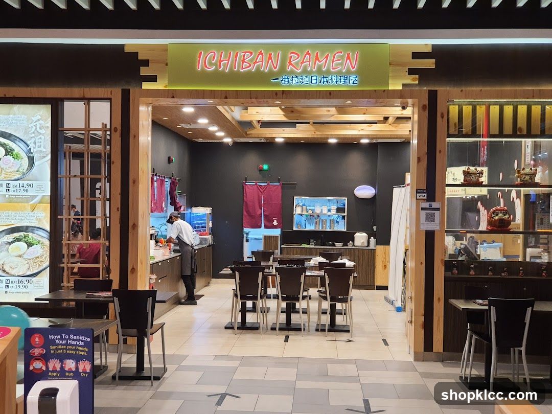 The exterior of a ramen restaurant with a prominent sign that reads "ICHIBAN RAMEN". The entrance is open, revealing the interior dining area and a staff member working behind the counter. To the left of the entrance, there are large posters displaying ramen dishes with prices.
