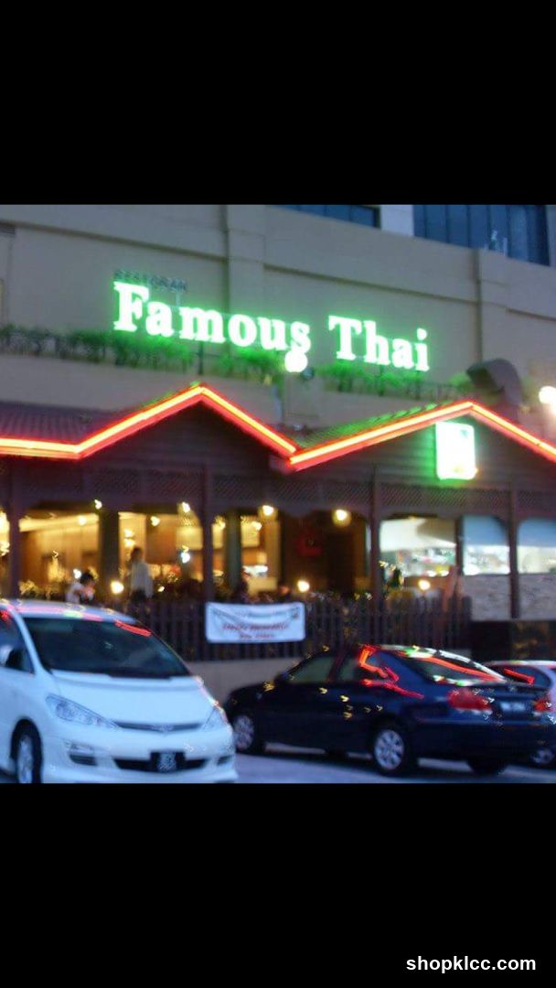The exterior of a restaurant called "Famous Thai" is visible. The restaurant has a prominent neon green sign with the name "Famous Thai" and a smaller sign above it that reads "RESTORAN". The building has a pitched roof with red and green neon trim. There are windows and an entrance visible, with some people inside. Cars are parked in front of the restaurant.