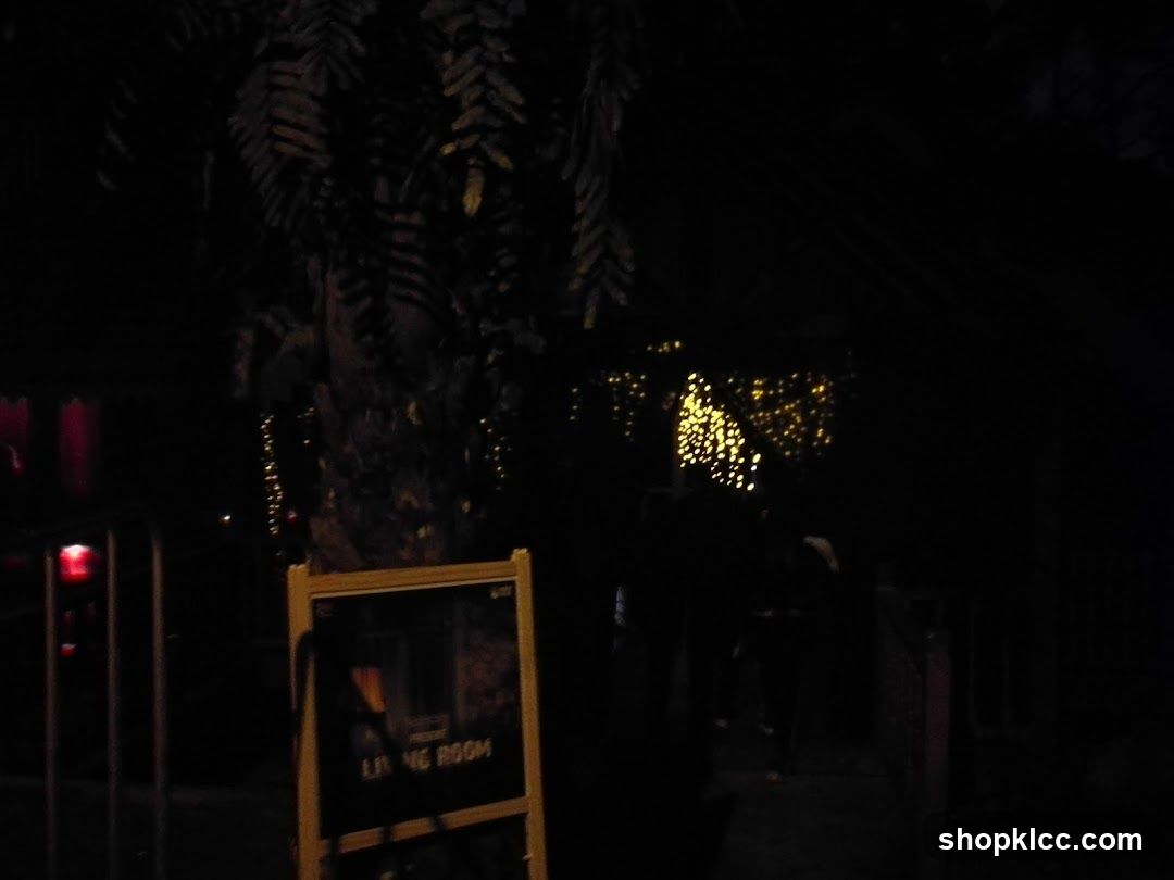 An A-frame sign with a dark background and white text that reads "LIVING ROOM" is visible in the foreground. The sign is illuminated by a yellow frame. In the background, there are out-of-focus string lights and foliage, suggesting an outdoor setting at night.