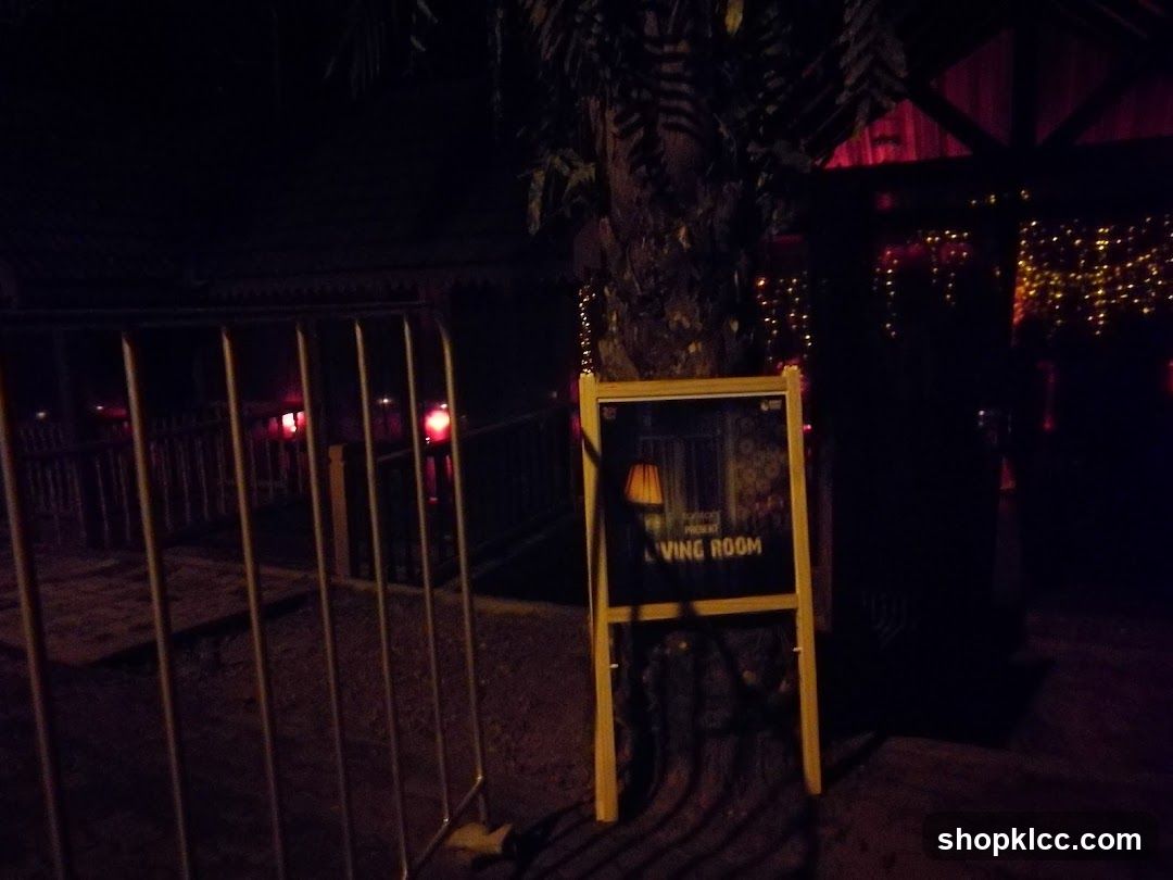 A sign on an easel displays an image of a lamp and the words "PRESENT LIVING ROOM". The sign is placed outdoors at night, with decorative lights visible in the background. A metal barrier is in the foreground.