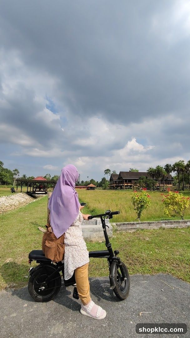 A woman wearing a purple hijab and a patterned tunic is sitting on a black electric scooter. She is holding a smartphone in her right hand. The scooter is parked on a paved path. In the background, there is a field of golden rice paddies, several traditional-style wooden buildings, and lush green trees under a cloudy sky.