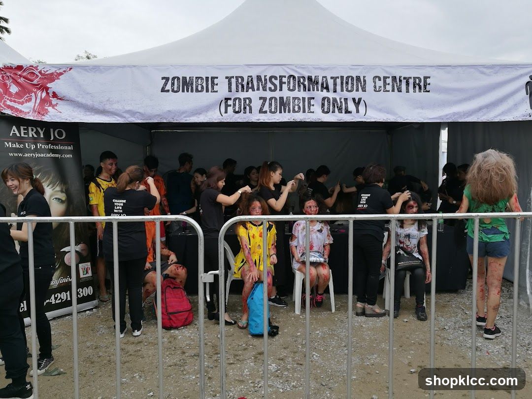 People are getting their makeup done to look like zombies under a tent. A banner reads "ZOMBIE TRANSFORMATION CENTRE (FOR ZOMBIE ONLY)". There is also a banner for "AERY JO Make Up Professional".