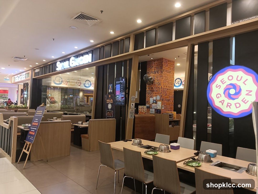 The exterior of a Seoul Garden restaurant, a Korean barbecue establishment. The restaurant features a modern facade with large windows and prominent signage. Inside, tables are set for dining, with grills integrated into the tabletops. A colorful wall mural inside displays various food items. A circular neon sign with the restaurant's logo is visible on the right side of the entrance.