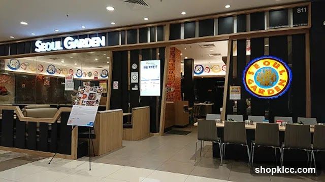 The exterior of a Seoul Garden restaurant with its prominent signage. The entrance features a modern design with wooden accents and glass panels. A large illuminated circular sign with the restaurant's name is visible on the right side. Inside, tables and chairs are set up, and a digital display shows "BUFFET". A stand with a menu or promotional material is placed near the entrance.