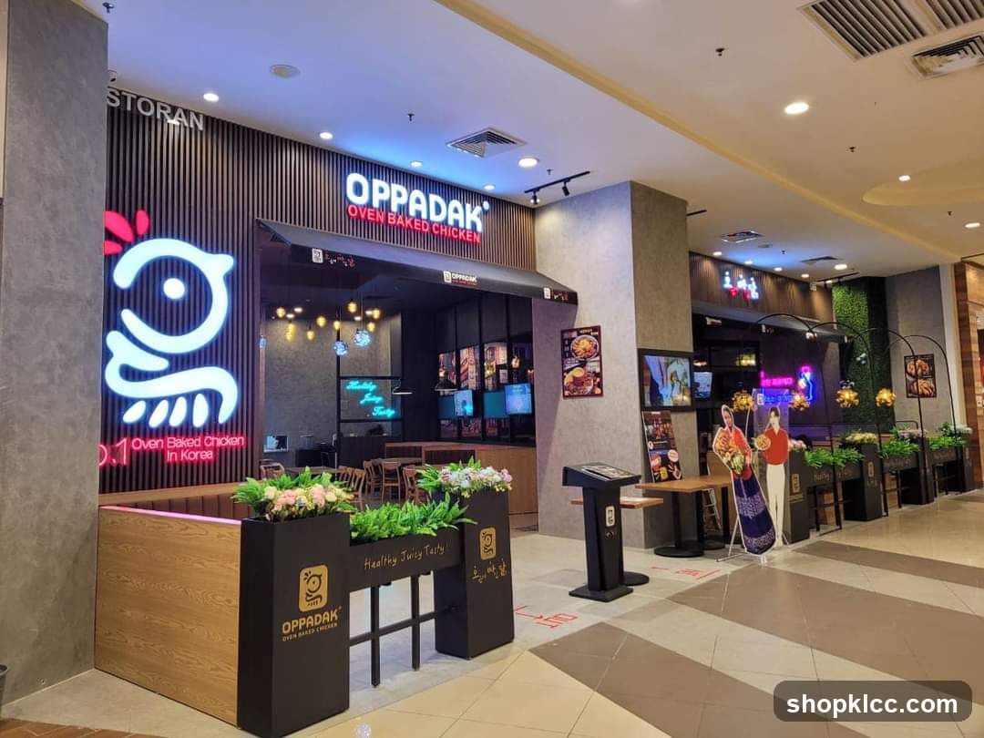 The exterior of an Oppadak restaurant, a Korean oven-baked chicken establishment. The storefront features a large, illuminated sign with the Oppadak logo, a stylized chicken, and the text "OPPADAK OVEN BAKED CHICKEN". Below the main sign, smaller text reads "Oven Baked Chicken In Korea". The entrance is partially visible, with seating and interior lights visible. Planters with greenery and flowers are placed in front of the seating area. A menu stand is positioned outside the entrance.
