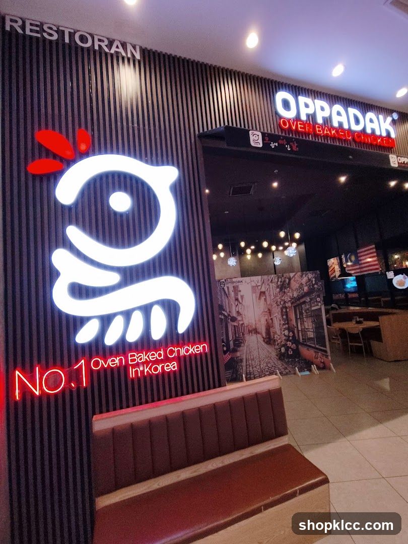 The exterior of a restaurant called Oppadak, specializing in oven-baked chicken. A large, illuminated logo of a stylized chicken is visible on the left, with the text "RESTORAN" above it. Below the chicken logo, neon red text reads "No.1 Oven Baked Chicken In Korea". To the right, the restaurant's name "OPPADAK" is displayed in large illuminated letters, with "OVEN BAKED CHICKEN" written below in red. A bench seating area is in the foreground. The interior of the restaurant, with tables and chairs, is visible through the entrance.