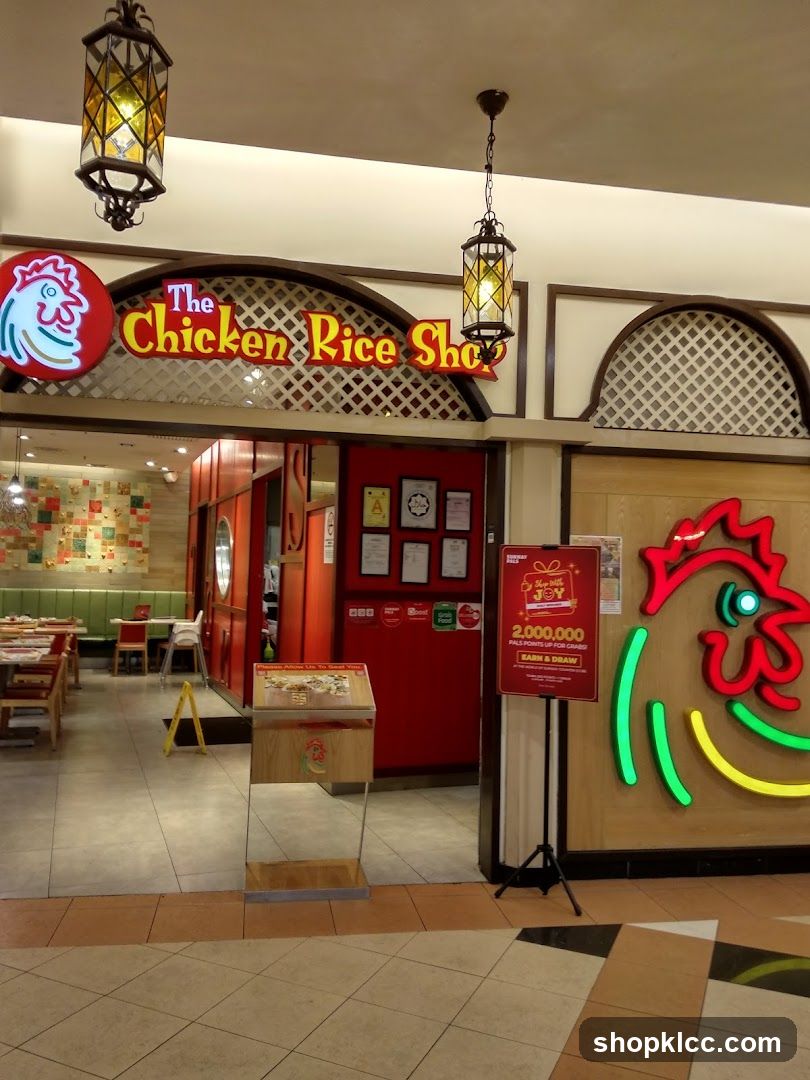 The exterior of a restaurant called "The Chicken Rice Shop". The entrance is visible with a red interior and tables and chairs set up inside. A large neon chicken sign is on the right side of the building. A promotional sign for "Shop with Joy" is displayed outside.
