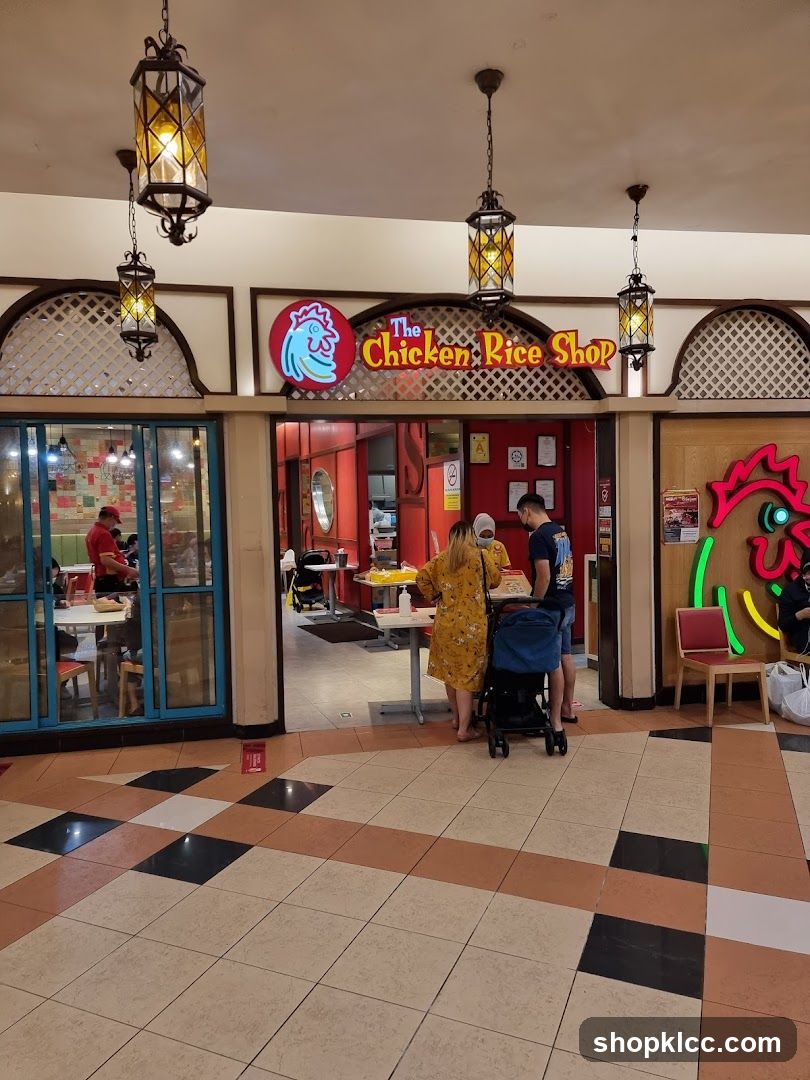 The exterior of a restaurant called "The Chicken Rice Shop". The entrance is open, and people are seen inside and at the counter. There are tables and chairs visible inside. The shop has a red and white color scheme with a prominent chicken logo.