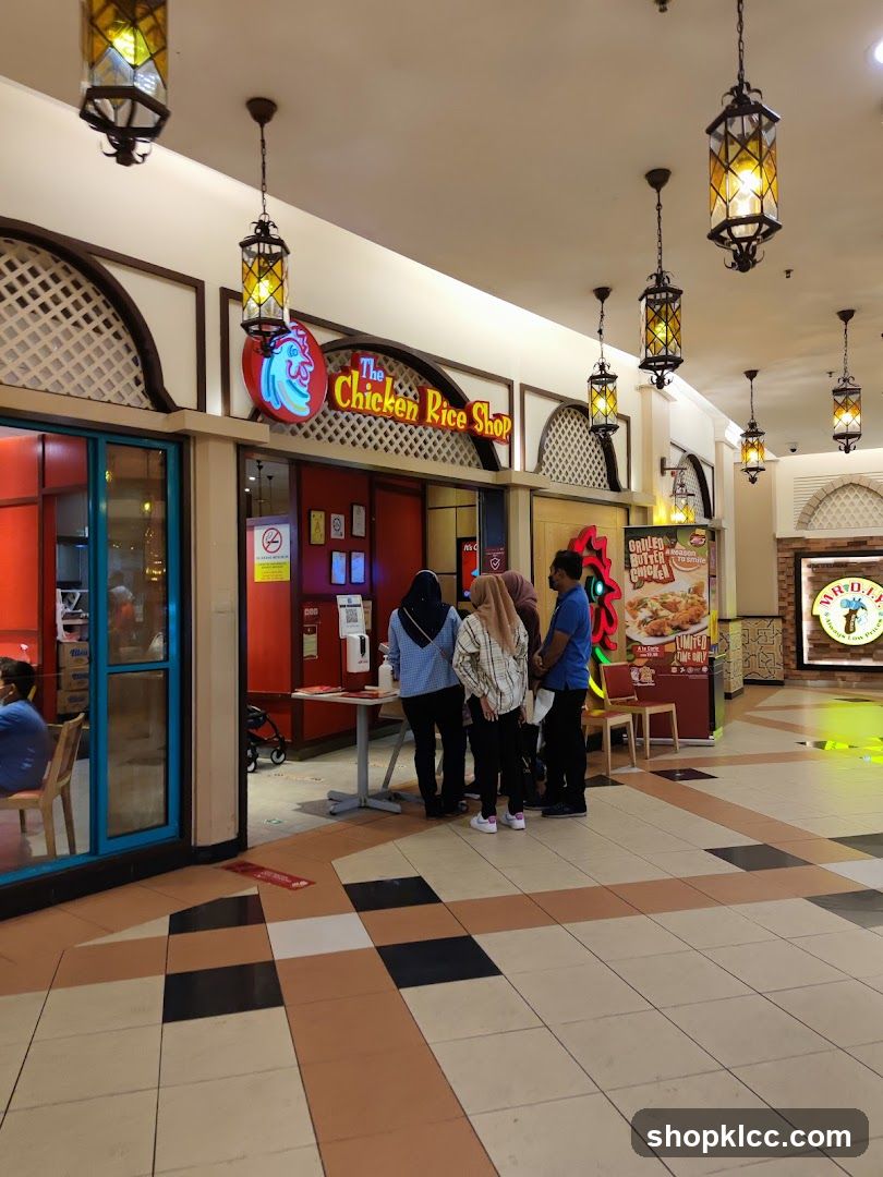 The exterior of a restaurant called "The Chicken Rice Shop" with a neon sign featuring a chicken logo. Several people are standing in front of the entrance, possibly in a queue. There is a promotional poster to the right of the entrance. The restaurant is located in a mall with decorative lanterns hanging from the ceiling.