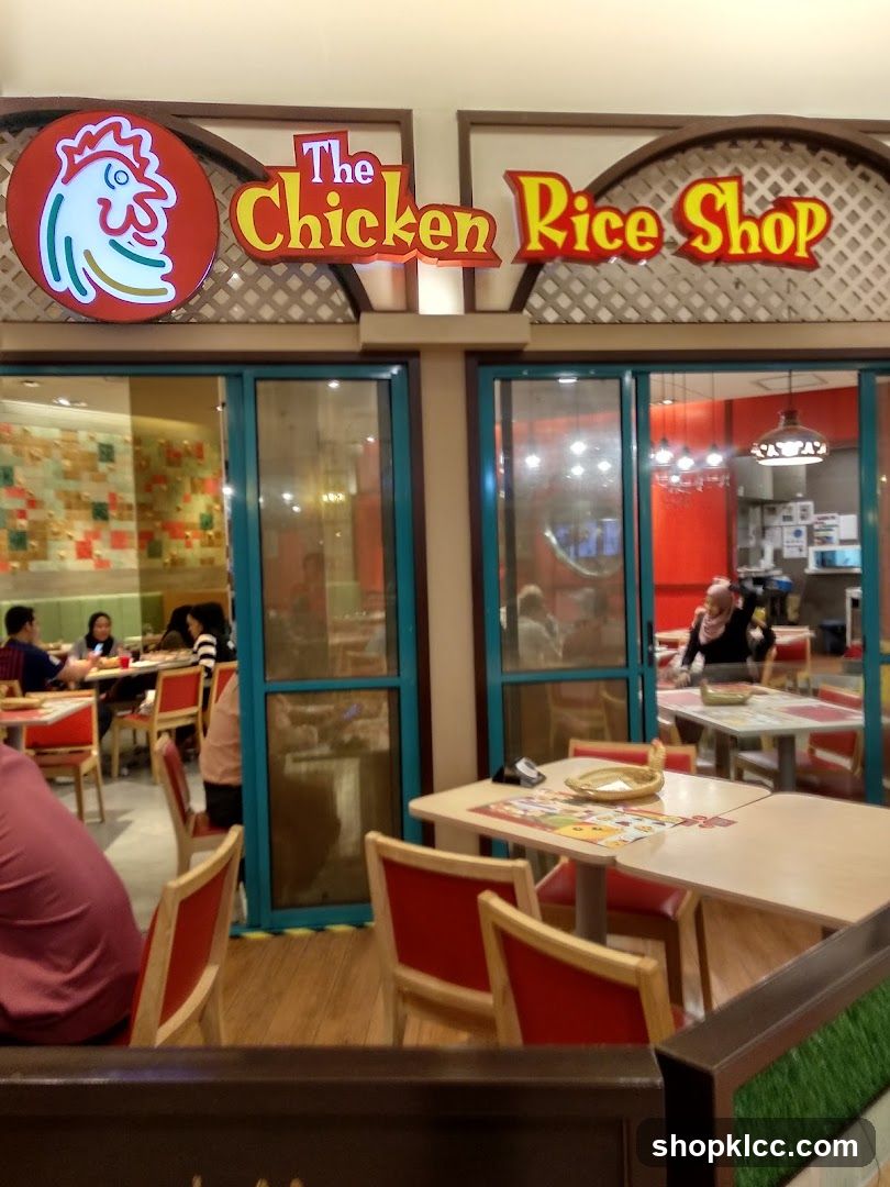 The exterior of a restaurant called The Chicken Rice Shop. The restaurant has large glass windows and doors, with red chairs and tables visible inside. The signage for the restaurant is prominently displayed above the entrance.