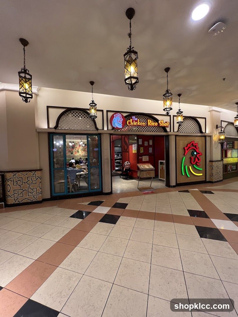 The exterior of a restaurant called "The Chicken Rice Shop". The entrance features a neon chicken sign and the restaurant's name is displayed above the doorway. Inside, diners can be seen at tables. The restaurant is decorated with patterned tiles and hanging lanterns.