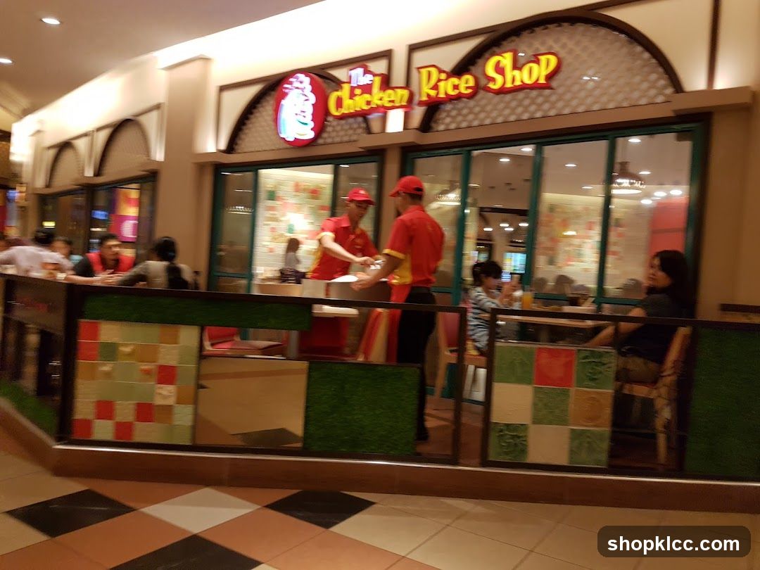 The exterior of a restaurant called "The Chicken Rice Shop" with a red and yellow sign above the entrance. People are seated inside and outside the restaurant.