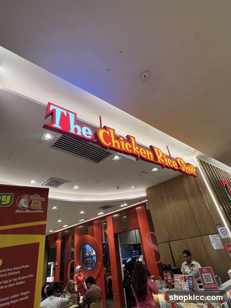A brightly lit interior of a restaurant with a prominent sign that reads "The Chicken Rice Shop". The sign is illuminated with red and blue neon lights. The restaurant has a modern design with wooden accents and red vertical stripes. Several customers are visible inside, some seated at tables and others queuing at the counter. A staff member is attending to customers at the counter.