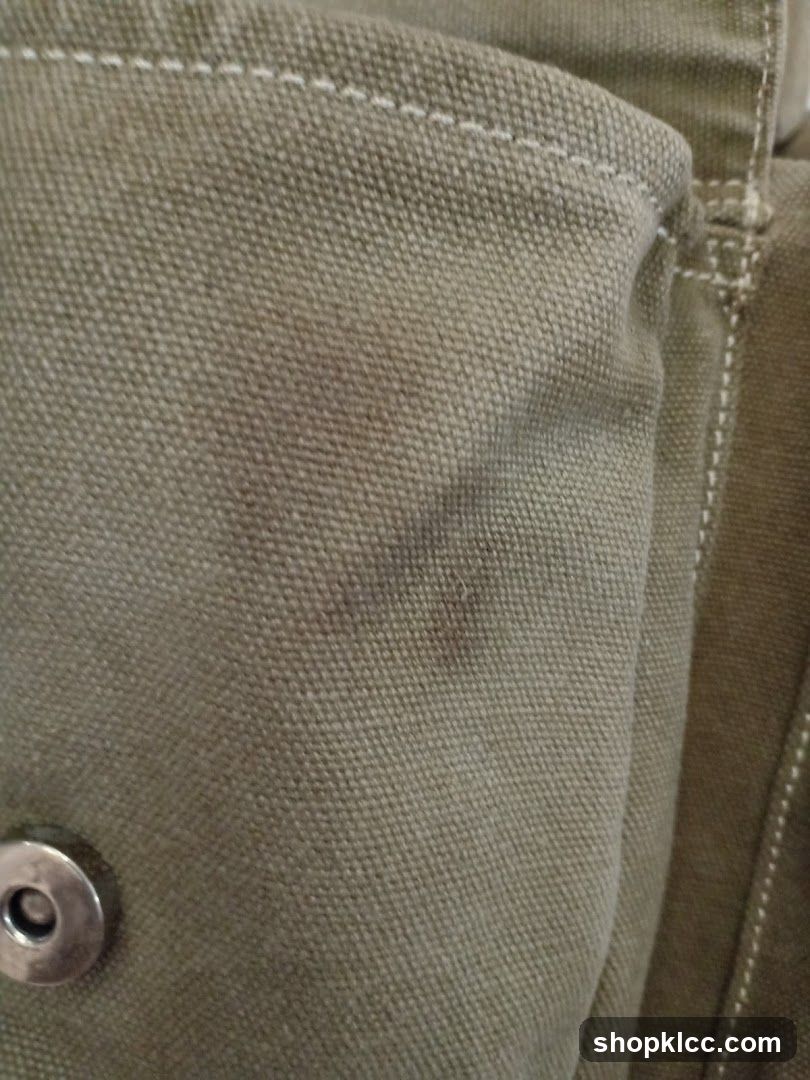 A close-up, detailed view of a textured, olive-green fabric, possibly canvas. White stitching is visible along a seam. A circular metal snap closure is also present on the left side of the image. The fabric appears to be part of a bag or garment.
