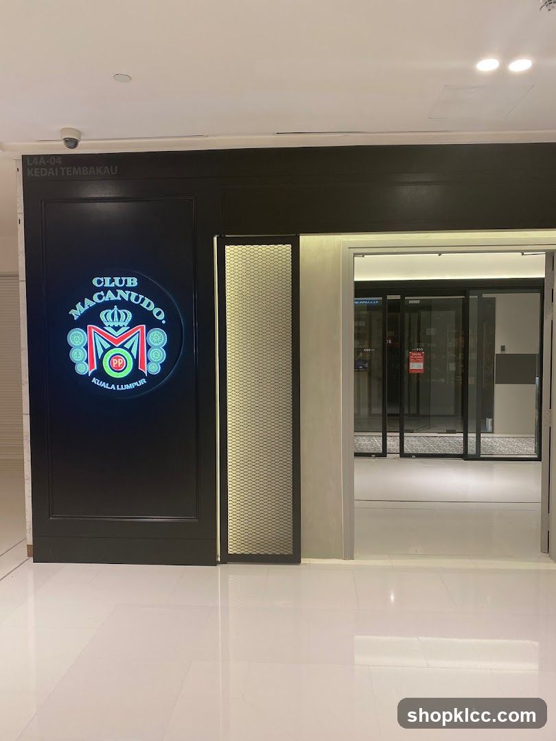 The exterior of Club Macanudo, a business located in Kuala Lumpur. The entrance features a prominent illuminated sign with the club's logo and name. To the right of the sign is a decorative panel, and further to the right are automatic glass doors leading into the establishment. Above the entrance, a sign indicates the unit number L4A-04 and the Malay phrase KEDAI TEMBAKAU.