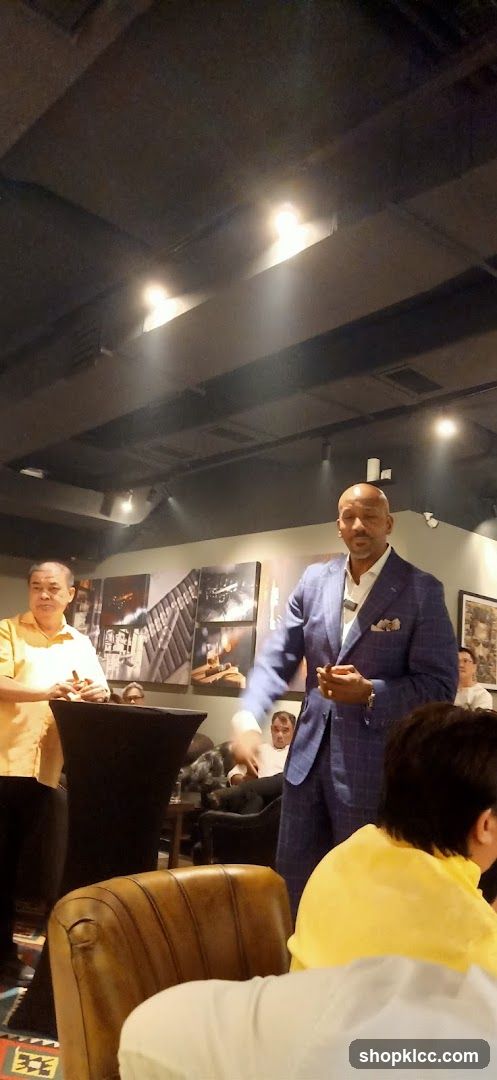 A man in a blue checkered suit is speaking to a group of people in a dimly lit room. He is holding a cigar. To his left, another man in a yellow shirt is also holding a cigar. The walls are decorated with framed pictures of what appear to be cigars and industrial scenes. Several people are seated in armchairs, and others are standing. The overall ambiance suggests a social gathering or event.