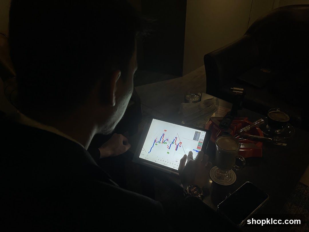 A man in a suit is looking at a tablet displaying stock market charts. The tablet is placed on a table with a drink, a phone, and an ashtray with cigars. The scene is dimly lit, suggesting a private or evening setting.