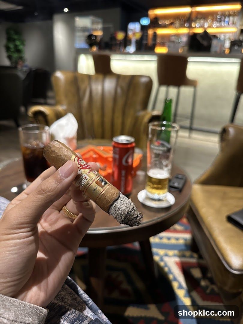A person holds a lit cigar in their hand, with a drink and a can of Coca-Cola on a table in front of them. In the background, a bar area with bottles and glasses is visible, along with leather armchairs and a patterned rug. The ambiance suggests a lounge or bar setting.