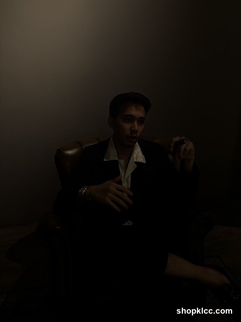 A man in a suit and open-collared shirt sits in a leather chair, holding a cigar. The lighting is dim, creating a dramatic atmosphere.