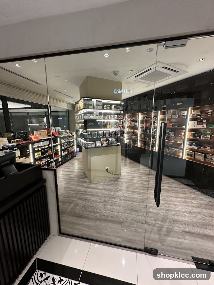 View through a glass door into a well-lit store. Shelves are stocked with numerous boxes, likely cigars or similar products. A central display case and a counter are visible. The floor has a wood-like tile pattern.