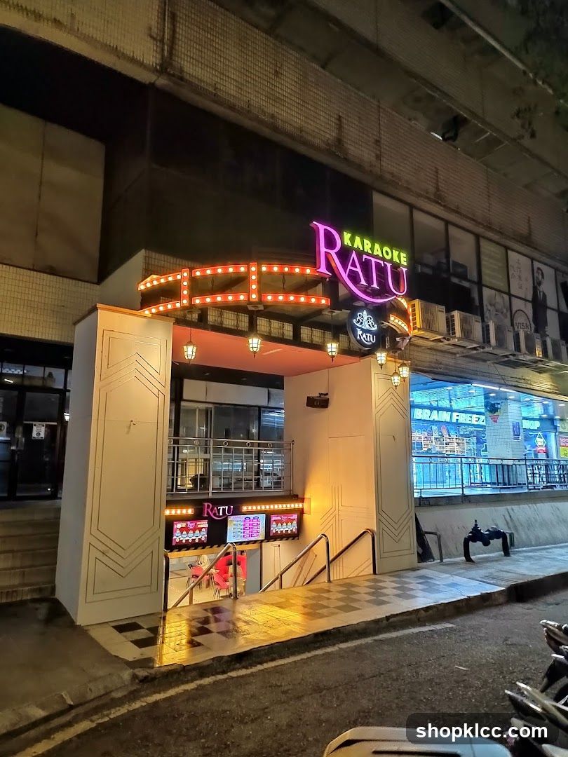 Night view of the exterior entrance to Karaoke Ratu, featuring a brightly lit neon sign with 'KARAOKE RATU' in pink and green letters. The building facade includes decorative light strips above the entrance, leading down a ramp with checkered flooring. A digital display board is visible near the entrance showing menu options or promotions. Adjacent storefronts are partially visible, including one labeled 'BRAIN FREEZ'.