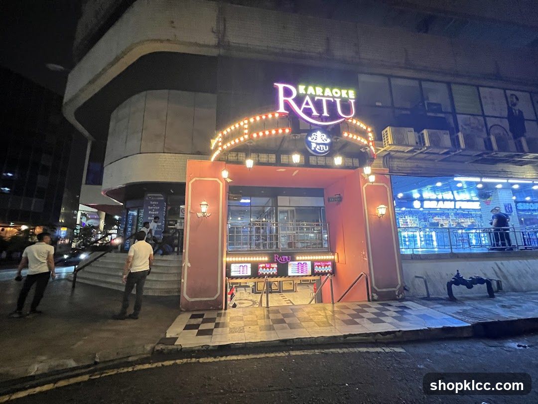 Night view of the exterior entrance to Karaoke Ratu, featuring illuminated signage with neon lights and decorative bulbs above a checkered floor entryway. Two men stand near the steps leading up to the building.
