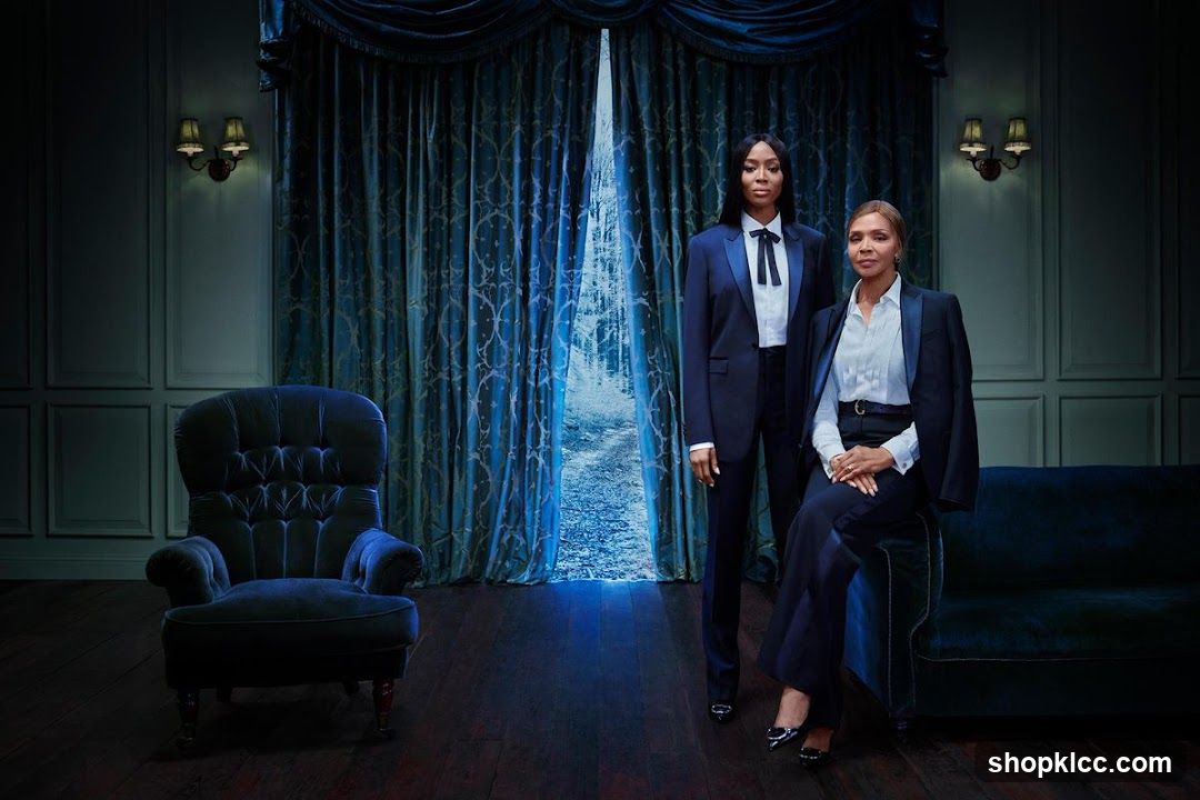 Two women in formal attire standing and sitting in a dimly lit room with blue curtains.