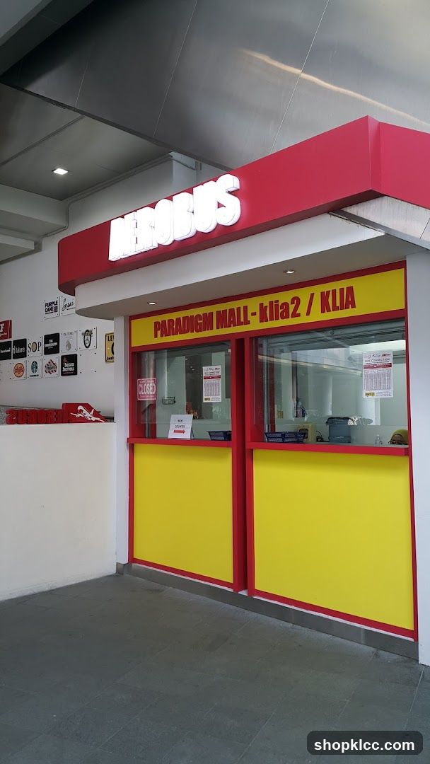 A bright red and yellow ticket counter with the word "AEROBUS" in large white illuminated letters above. The counter has a yellow lower section and a red frame. Above the windows, a red sign reads "PARADIGM MALL-klia2 / KLIA". A "CLOSED" sign is visible on the left window, and a "NEXT COUNTER" sign is on the right. Inside, bus schedules are posted on the windows.