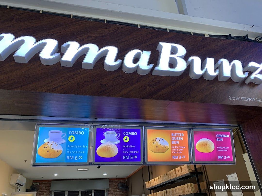 Exterior view of a bakery with illuminated signage reading 'MmaBunz' on a dark wood panel. Below the sign, four digital menu boards display combo options and individual bun offerings with prices in Malaysian Ringgit (RM). The interior is partially visible, showing brick walls and shelving units.