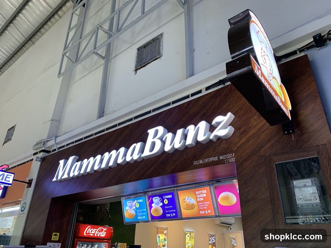 Exterior view of a bakery with illuminated 'MammaBunz' sign on a dark wood panel facade. Above the entrance, there is an LED display showing combo options and product images like Butter Queen Bun. A Coca-Cola refrigerator is visible inside the shop. The building has industrial-style ceiling and metal framework.