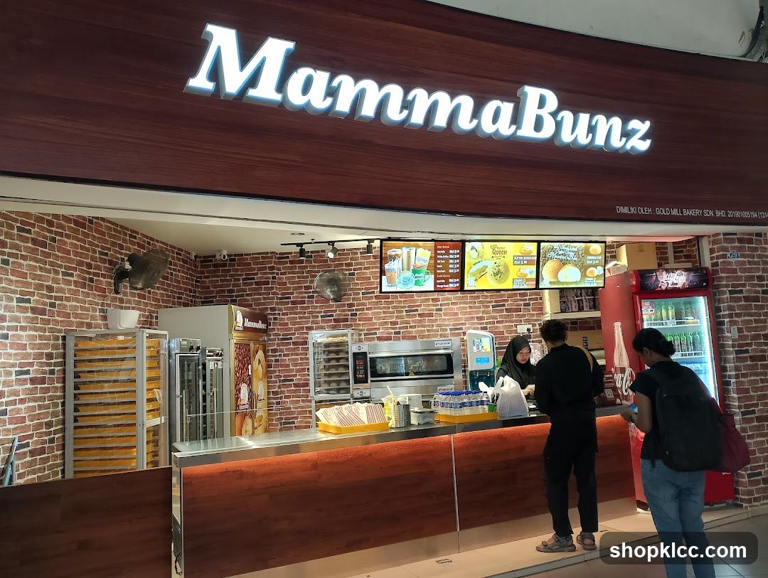 Storefront with illuminated 'MammaBunz' sign above a brick-walled counter area. Customers are at the service counter; behind it, there's an oven, refrigerated display units, and digital menu boards showing food items and prices.