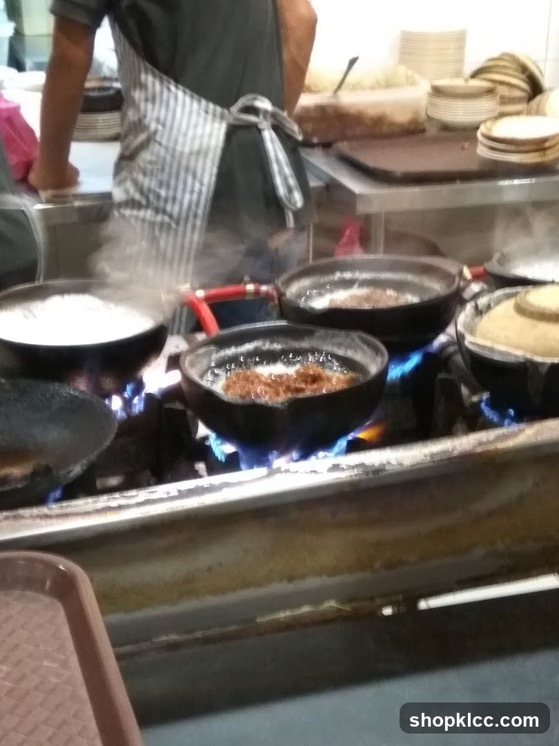 A chef in a striped apron stands behind a stove with multiple pots on gas burners emitting steam. The kitchen is equipped with stacked plates and bowls in the background. The environment suggests an active food preparation area.