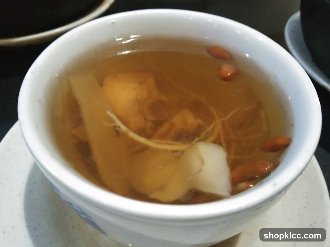 A white ceramic bowl filled with a light brown broth containing ingredients such as ginseng roots, goji berries, and possibly slices of ginger or other herbs. The bowl rests on a speckled saucer. Background shows partial view of other dishes or plates on a dark surface.