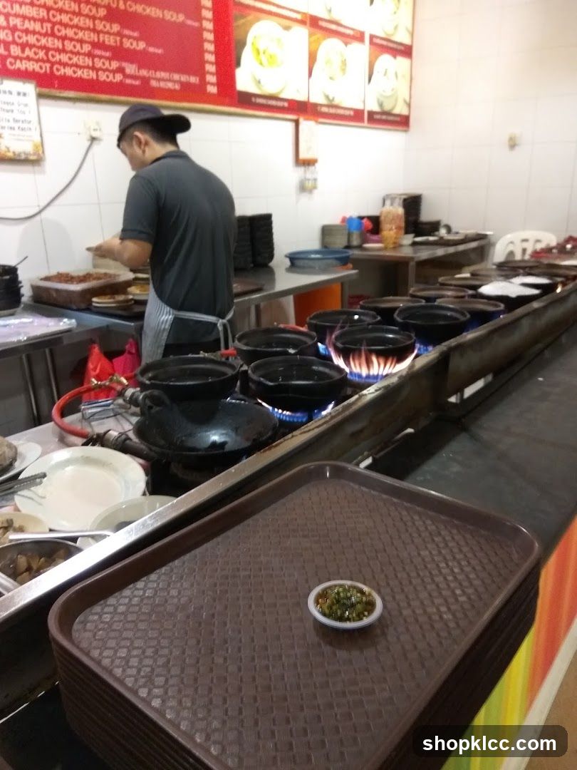 Interior view of a food preparation area showing a staff member in a dark shirt and cap working behind a counter. Multiple black pots are on gas burners with blue flames. A brown tray holds a small bowl of green chili. In the background, a red menu board lists soup options like Cucumber Chicken Soup and Peanut Chicken Feet Soup. The walls are tiled white, and there are stacks of plates and bowls nearby.
