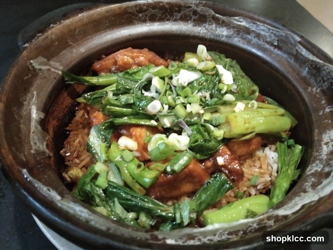 A close-up shot of a dish served in a rustic clay pot, featuring rice topped with glazed meat pieces, green vegetables, and chopped scallions.