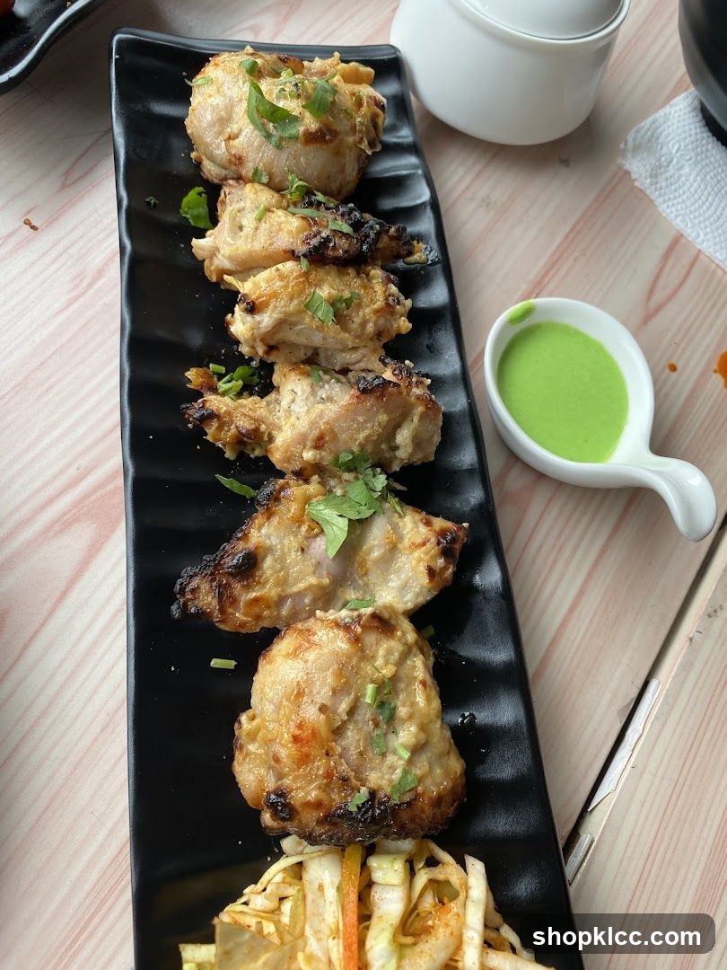 Grilled chicken pieces garnished with herbs served on a black rectangular plate alongside a side of coleslaw and a small bowl of green sauce. The setting is a light wooden table with a white ceramic cup and napkin nearby.