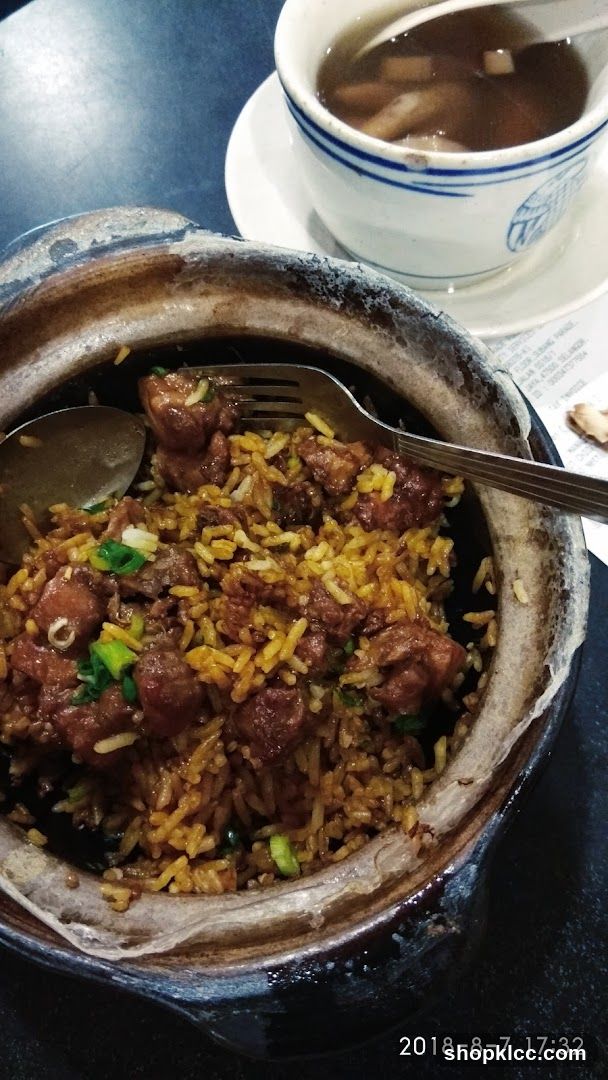 A close-up shot of a clay pot containing rice mixed with meat pieces and green onions, accompanied by a cup of soup on a saucer. The setting appears to be indoors at a dining table. A timestamp is visible in the corner.