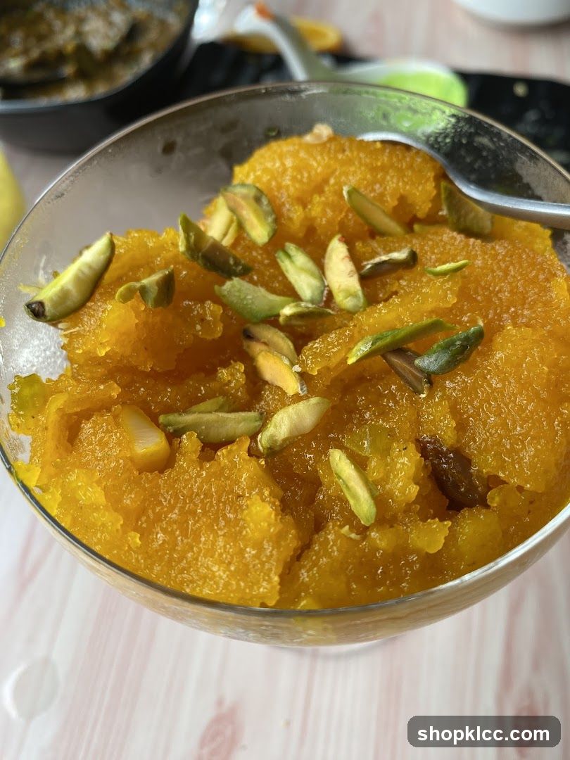 A close-up shot of a golden-orange textured dessert topped with sliced pistachios, served in a clear glass bowl with a spoon resting inside. The background shows part of another dish and a light-colored wooden table surface.