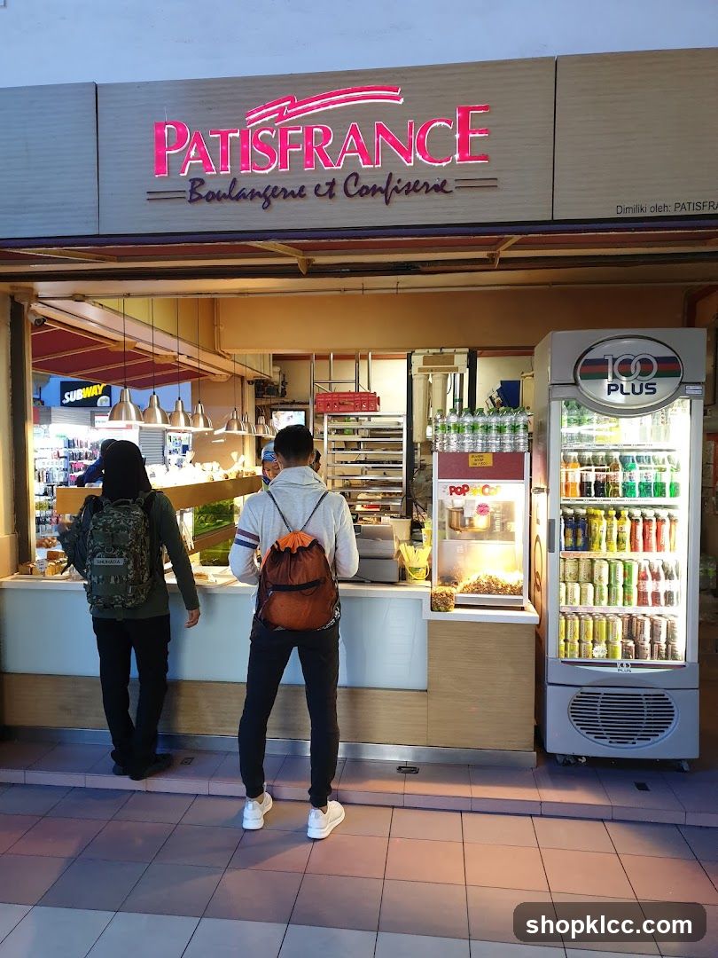 Storefront with pink neon sign reading 'PATISFRANCE Boulangerie et Confiserie' above counter. Customers are at the counter; a 100 PLUS beverage refrigerator is to the right. Interior lighting illuminates pastries and snacks on display. A SUBWAY sign is partially visible in background.