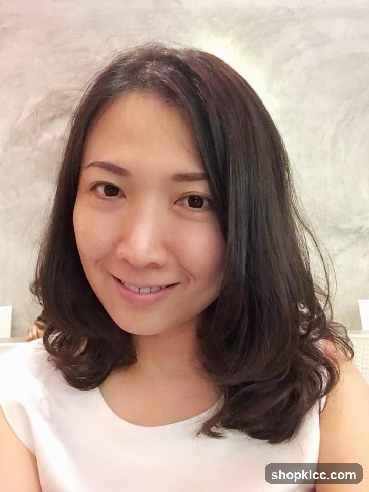 Portrait of a woman with shoulder-length dark hair smiling gently at the camera, wearing a white sleeveless top against a light textured wall background.