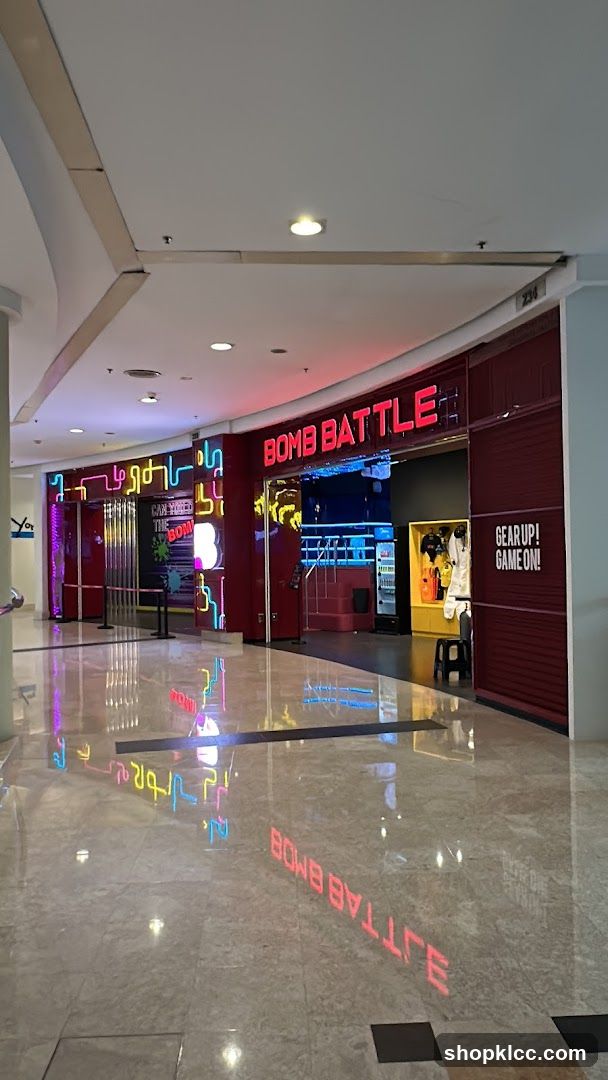 The exterior of a business called "Bomb Battle" is shown. The entrance features bright neon lights and a large red sign with the business name. To the right of the entrance, there is a display of white and yellow gear, with a sign that reads "GEAR UP! GAME ON!". The floor reflects the neon lights and the business name.