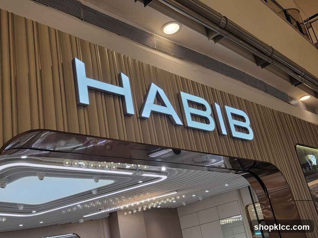A close-up shot of the exterior signage of a store. The name "HABIB" is displayed in large, illuminated white letters against a textured, light brown wooden panel wall. Below the signage, a curved, reflective metallic frame outlines the entrance area, which features modern lighting and a glimpse of the interior.