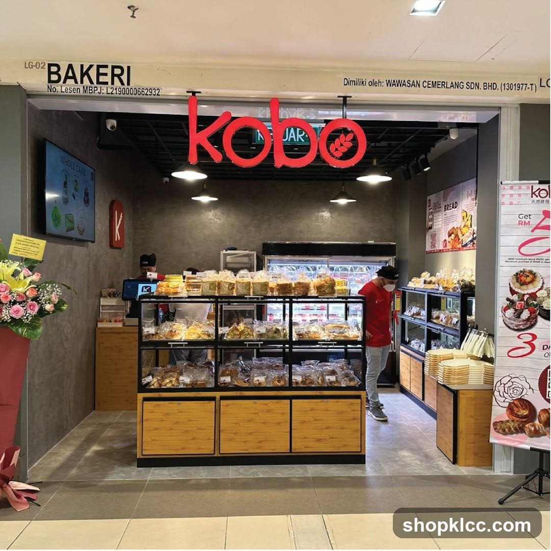 The exterior of a bakery with a large red "kobo" sign above the entrance. Display cases filled with bread and pastries are visible inside. A person in a red shirt and mask is standing inside the shop. Promotional posters are displayed to the right.