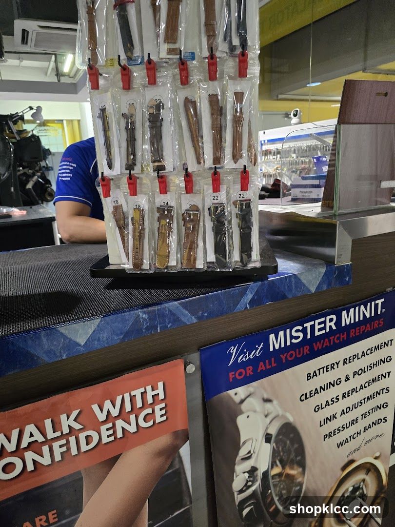 A display of various watch straps in plastic packaging, hanging from a rack. Below the watch straps are two promotional posters. One poster reads "WALK WITH CONFIDENCE" and the other, for "MISTER MINIT", lists various watch repair services including battery replacement, cleaning & polishing, glass replacement, link adjustments, pressure testing, and watch bands. A person in a blue shirt is visible behind the counter.